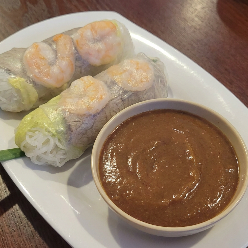 Three shrimp summer rolls with vermicelli and vegetables served alongside peanut dipping sauce on a white plate.