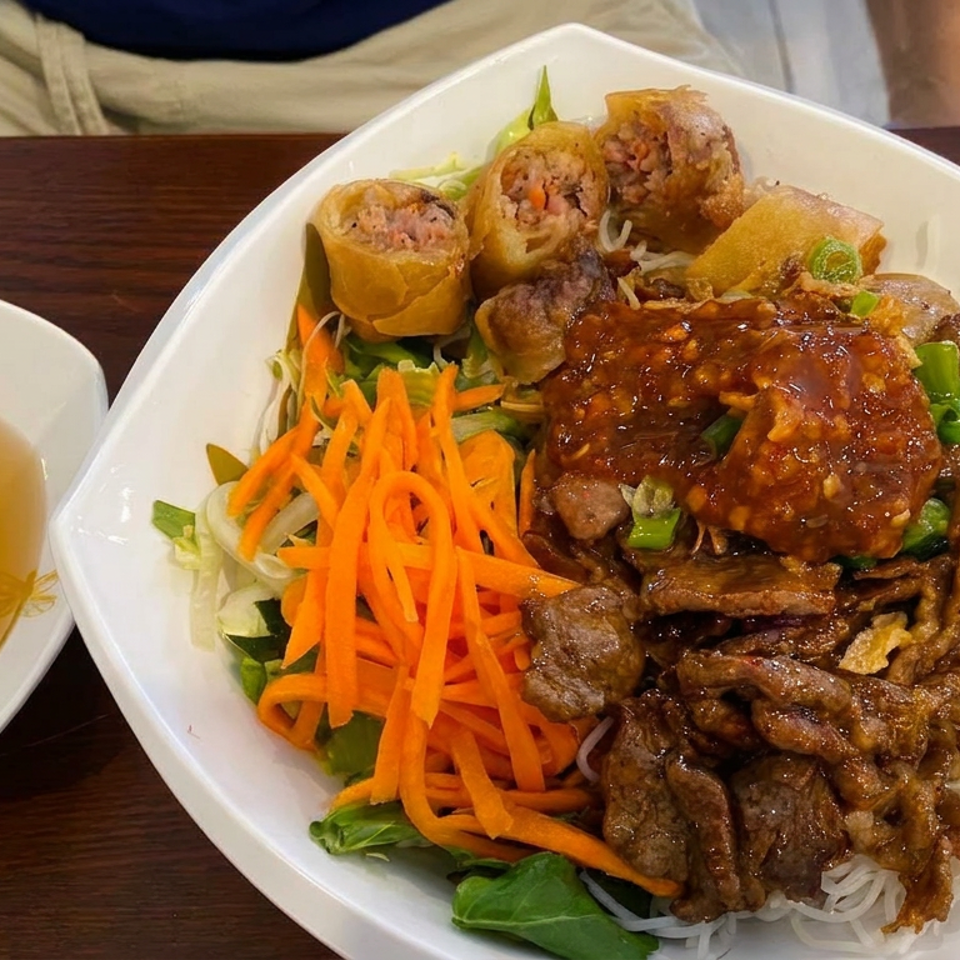 Vietnamese vermicelli bowl topped with grilled beef, fried spring rolls, shredded carrots, greens, and sauce, served with dipping broth.